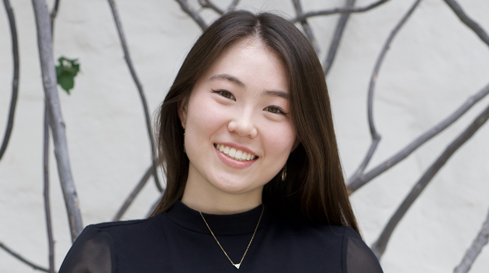 Portrait of a current Gannon Scholar with long dark hair, smiling, wearing a black top and a delicate necklace, standing in front of a light wall with tree branches in the background.